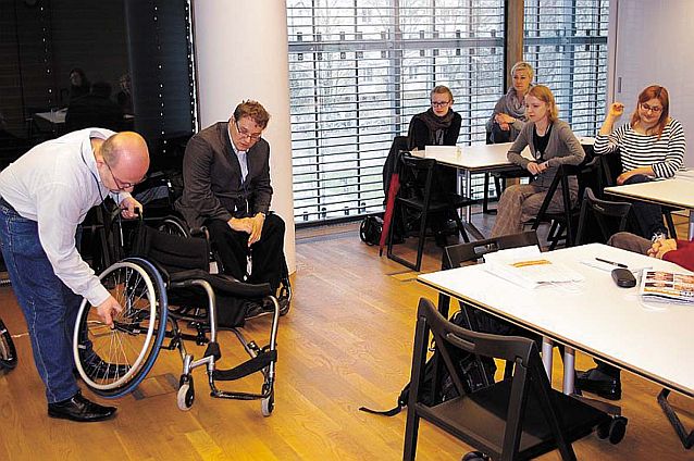 Dwóch mężczyzn prezentuje grupie obserwatorów, jak rozłożyć wózek inwalidzki