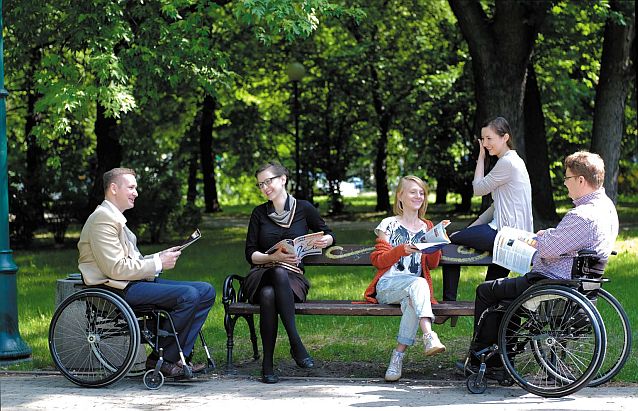 Dwie osoby siedzące na wózkach i trzy siedzące na ławce w parku