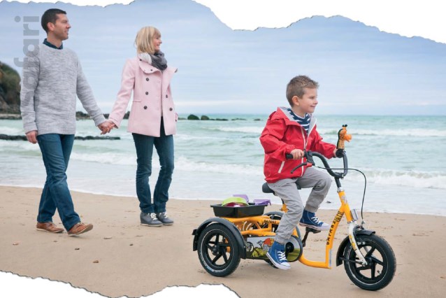 dziecko jedzie po plaży na trójkołowym rowerku, za nim idą rodzice 