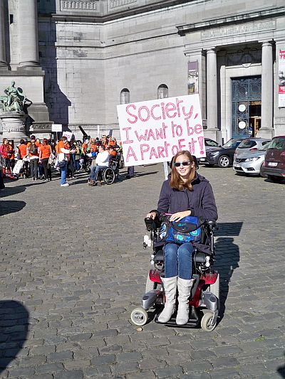 Młoda kobieta na wózku elektrycznym z transparentem z napisem: Society, I want to be a part of it!