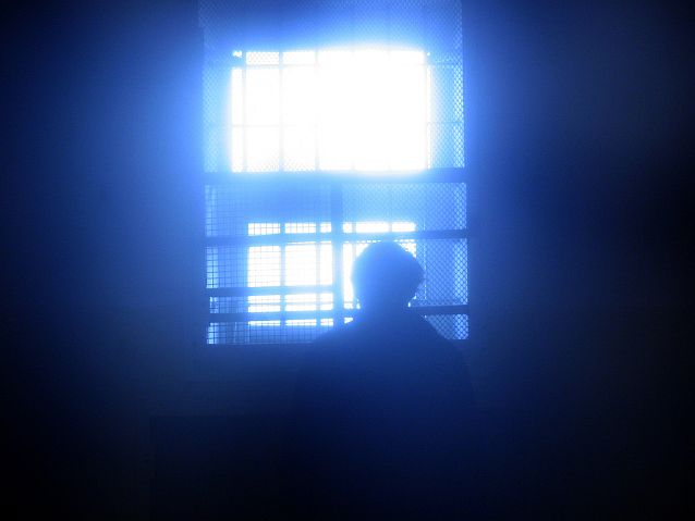 Człowiek stoi przed zakratowanym oknem, przez które wpada światło