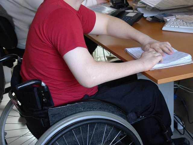 fragment osoby siedzącej na wózku inwalidzkim, która trzyma dłonie na dokumentach, leżących tuż przy klawiaturze komputera