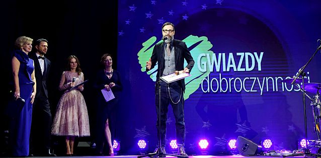 Szymon Majewski przy mikrofonie na scenie podczas Gali, obok stoją prowadzący