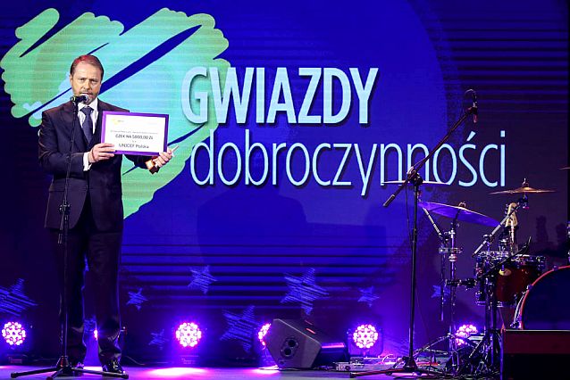 Artur Żmijewski na scenie przy mikrofonie z czekiem dla UNICEF Polska w rękach. W tle napis: Gwiazdy Dobroczynności