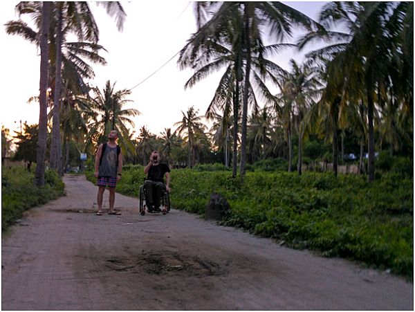 Dwaj mężczyźni, w tym jeden na wózku, na piaszczystej drodze, wokół palmy