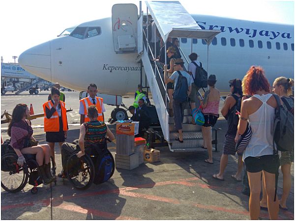 Dwie osoby na wózkach stoją przed samolotem. Inni wchodzą do niego po schodkach