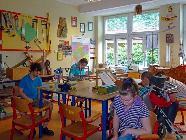 dorosłe osoby z niepełnosprawnością siedzą przy ogromnym biurku w pracowni
