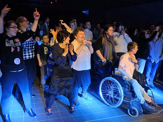 Tańczący ludzie z rękami w górze, wśród nich osoba na wózku