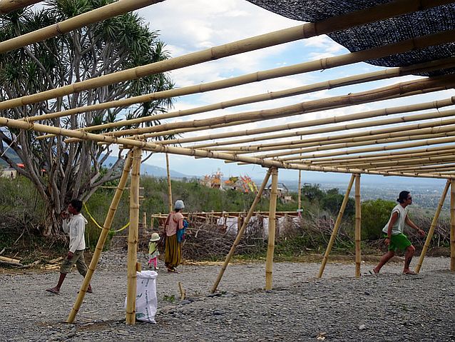 bambusowa konstrukcja, obok której po ziemi wyłożonej kamykami przechadzają się ludzie