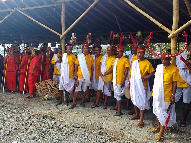 grupka kobiet ubranych na czerwono stoi z lewej strony, z prawej grupa mężczyzn w żółtych koszulach, białych spodniach i tradycyjnych ozdobach na głowach