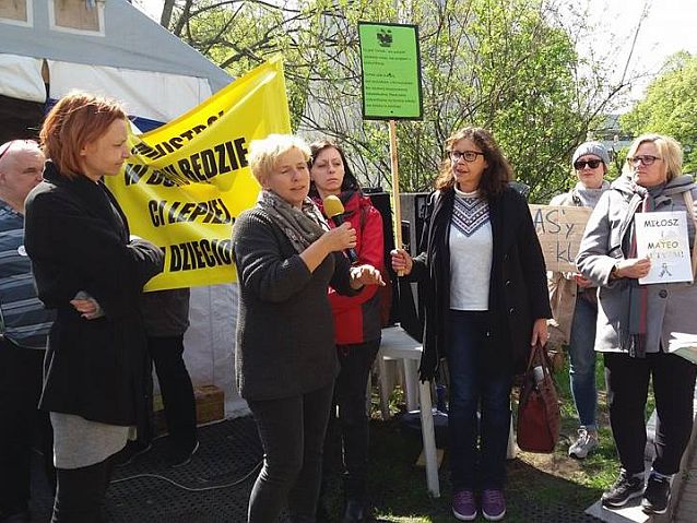 Kilka kobiet stoi pod żółtym transparentem. Jedna z nich mówi do mikrofonu