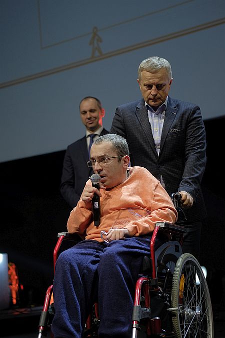 Do mikrofonu mówi siedzący na wózku Piotr Lewandowski, za nim stoi asystent