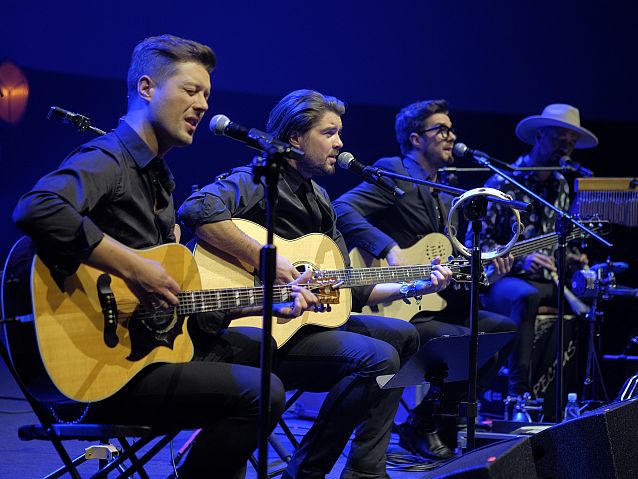 Zespół czterech mężczyzn, trzech siedzi grając na gitarach i śpiewając, czwarty gra na instrumentach perkusyjnych