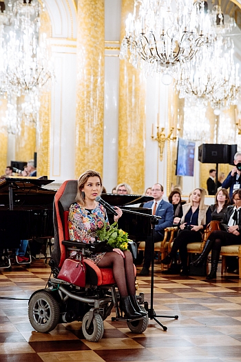 Agata Roczniak na wózku elektrycznym przemawia do mikrofonu w eleganckiej sali, w tle publiczność
