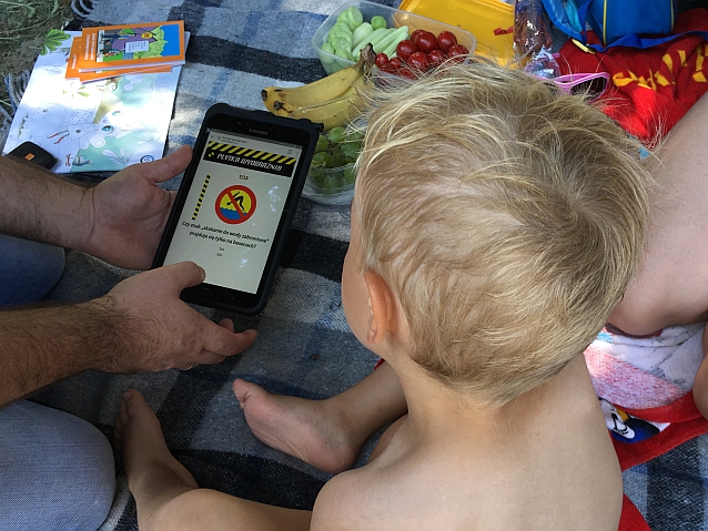 Siedzące na kocu dziecko patrzy w ekran smartfona, na którym wyświetla się pytanie z quizu dot. płytkiej wyobraźni