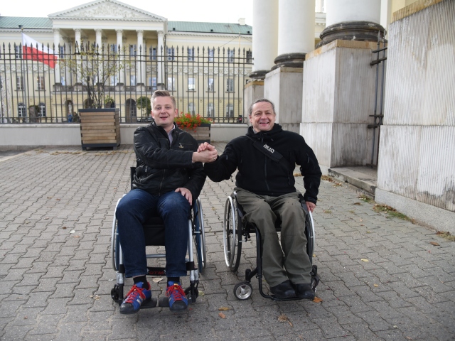 Jakub Stefaniak i mężczyzna z włosami związanymi w kucyk znajdują się na wózkach, uśmiechnięci podają sobie dłonie. Są przed bramą ratusza