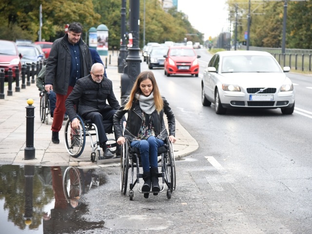 Przejazd przez ulicę z dużą kałużą. Pierwsza porusza się na wózku młoda kobieta, za nią przejeżdża przez krawężnik Sławomir Antonik, tuż za nim asekurant