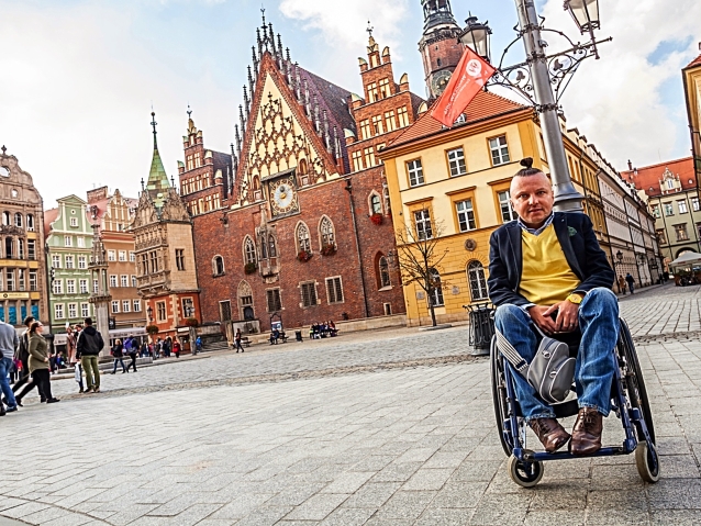 Bartłomiej Skrzyński na wózku pozuje do zdjęcia na wrocławskim Starym Mieście, w tle zabytkowe kamienice