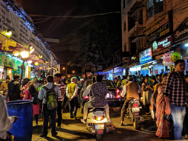 Noc w miasteczku indyjskim. Tłumy osób na ulicy, między pieszymi przejeżdża auto, za nim dwa motory.