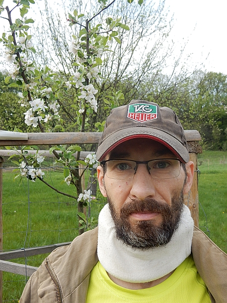 Arkadiusz Turoń w czapce bejsbolówce i kołnierzu ortopedycznym, w tle drzewko z rozkwitającymi białymi kwiatami