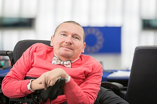 Marek Plura siedzi na wózku elektrycznym, w tle sala plenarna z flagą Unii Europejskiej
