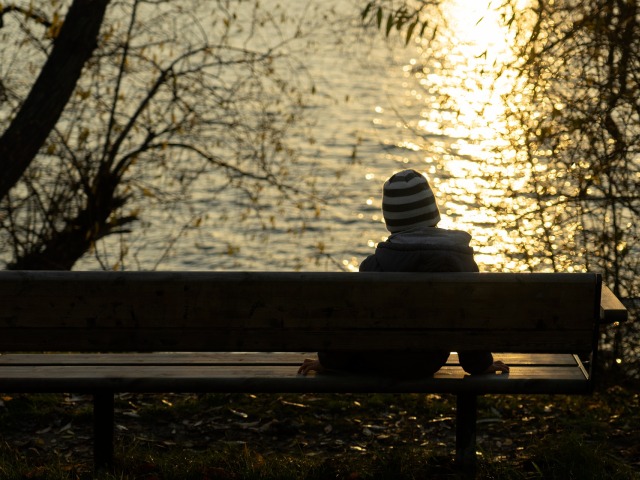 samotny kilkuletni chłopiec w czapce i kurtce siedzi tyłem do aparatu na ławce nad wodą