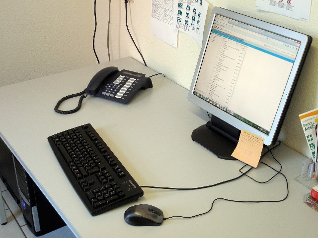 komputer stoi na biurku leży klawiatura i mysz obok stoi telefon stacjonarny