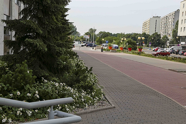 Kilkadziesiąt metrów pustego chodnika, dalej widać szlaban i parking z samochodami