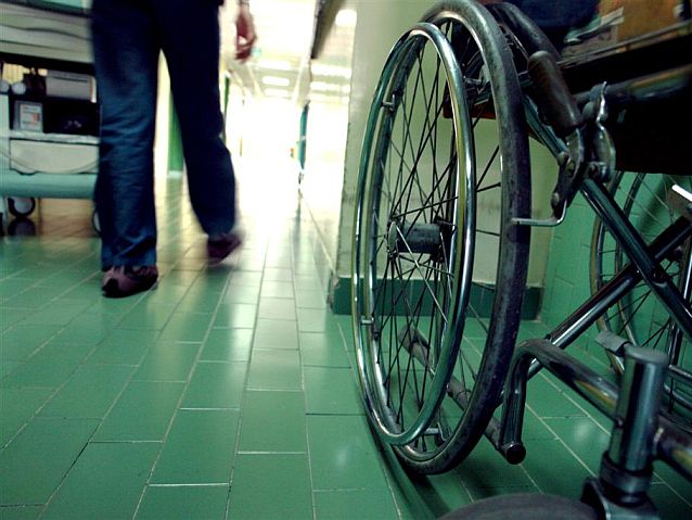 Wózek stoi na korytarzu przychodni lub szpitala, obok przechodzi człowiek