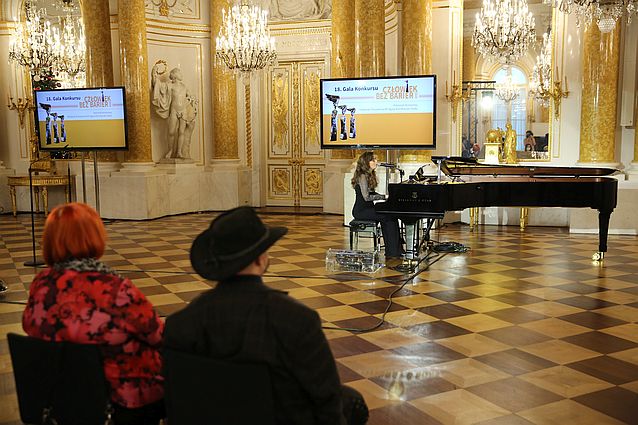 Kobieta i mężczyzna w wytwornej sali siedzą na krzesłach, słuchając występu pianistki, grającej na fortepianie