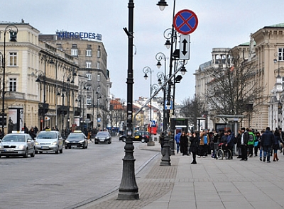 Widok na jezdnię i chodnik ul. Krakowskie Przedmieście. Na środku zamocowany do latarni znak drogowy