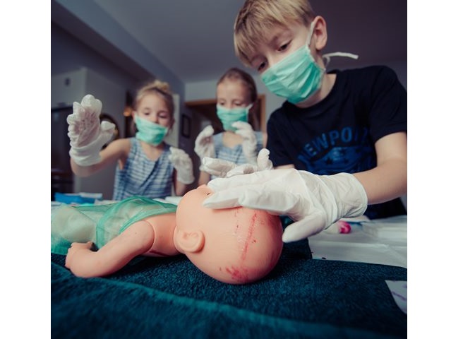 trójka dzieci w maseczkach pochyla się nad manekinem dziecka, chłopiec dotyka go rękami