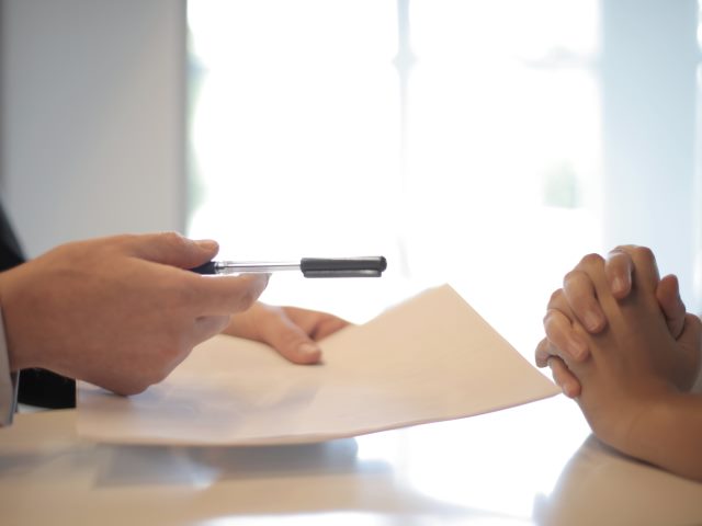 ręce męskie trzymają dokumenty i długopis nad stołem ręce damskie naprzeciwko założone jedna w drugą