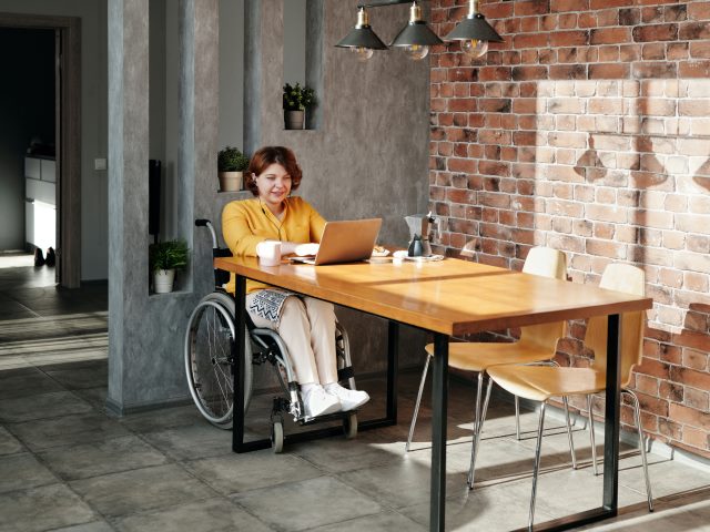 kobieta na wózku siedzi w słuchawkach w uszach na wózku przy stole na przy laptopie, jest uśmiechnięta