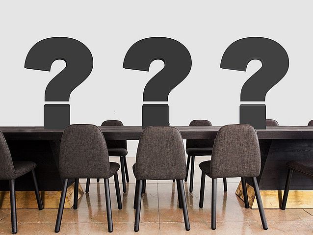 Grafika przedstawia trzy duże znaki zapytania stojące na stole otoczonym pustymi krzesłami