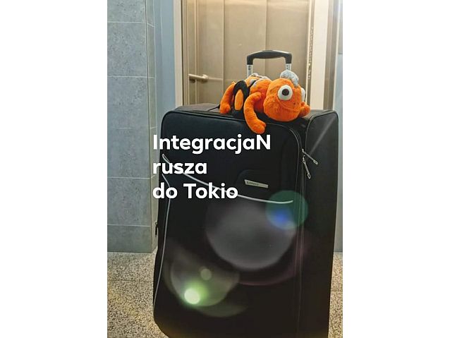 Na walizce siedzi pomarańczowa maskotka kameleona na wózku. Napis brzmi: IntegracjaN rusza do Tokio