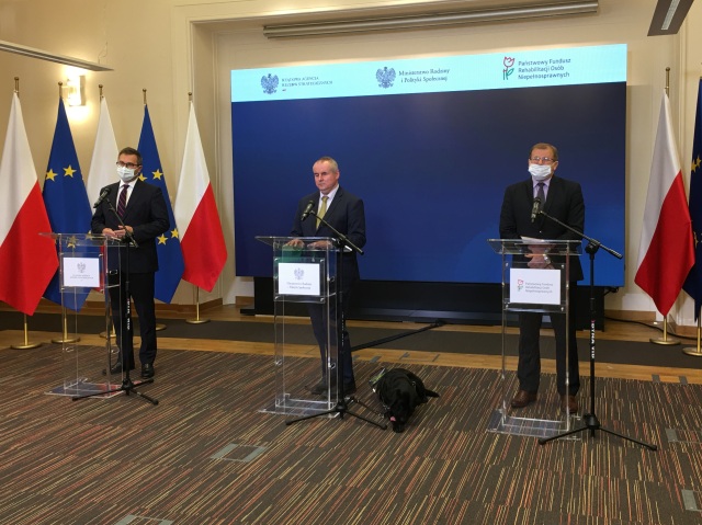 Konferencja. W sali, na tle flag Polski i Unii Europejskiej od lewej przy mikrofonach stoją: Michał Kuczmierowski, prezes Rządowej Agencji Rezerw Materiałowych, następnie pełnomocnik Paweł Wdówik i Tomasz Maruszewski