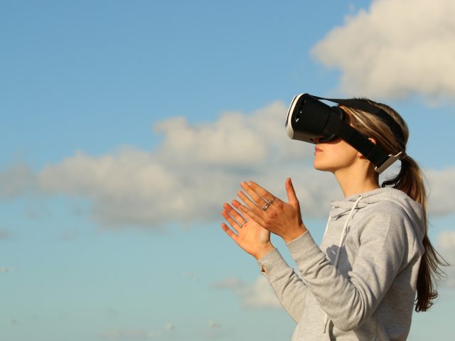 młoda dziewczyna w okularach VR wyciągnięte ręce do przodu na tle błękitnego nieba