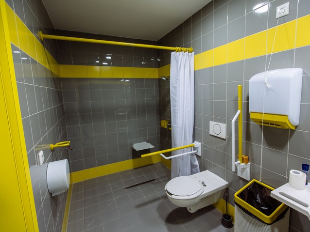 Wnętrze dostosowanej łazienki. Po prawej umywalka i muszla ustępowa, na końcu obszerny otwarty prysznic z krzesełkiem