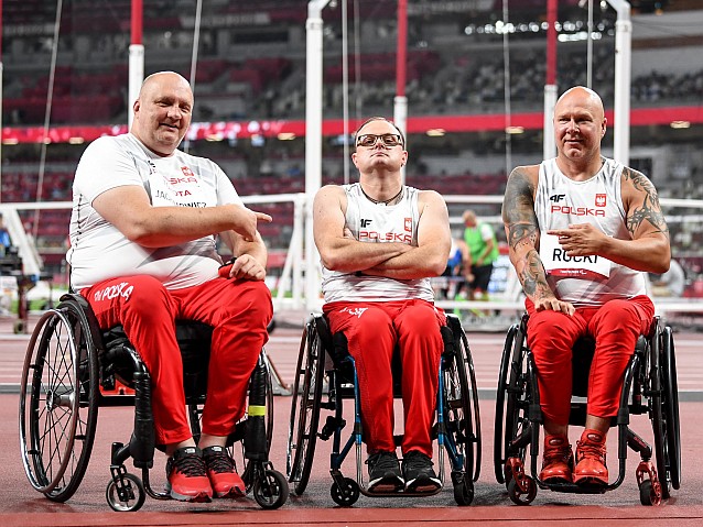 Trzech polskich zawodników na wózkach na stadionie lekkoatletycznym. Piotr Kosewicz w środku siedzi z założonymi rękami. Dwóch zawodników po bokach wskazuje na niego.