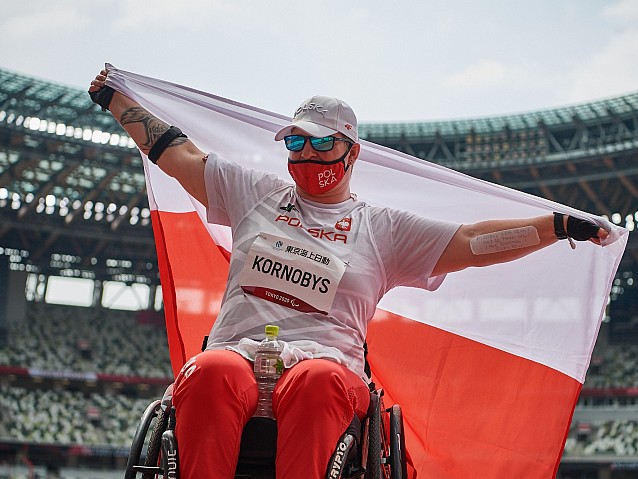 Lucyna Kornobys siedzi na wózku na tle stadionu, za plecami rozciąga flagę Polski