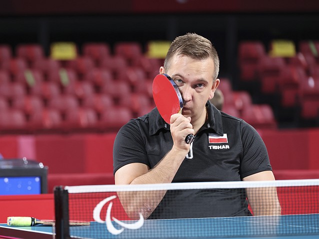 Siedzący na wózku przy stole do tenisa podłamany Rafał Czuper przykłada do policzka rakietkę