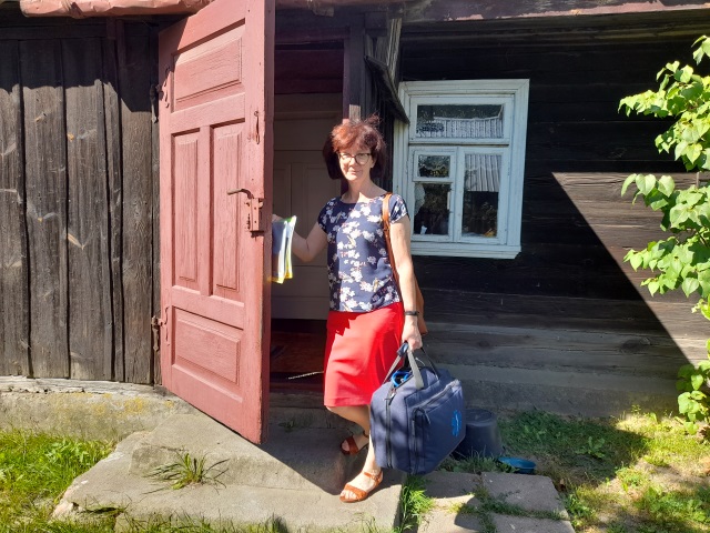 Doktor Ewa Stankiewicz stoi przy drzwiach drewnianego domu. W dłoni ma torbę lekarską