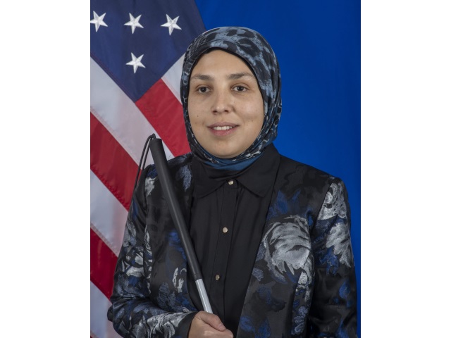 Kobieta z biała laską i w hidżabie stoi na tle amerykańskiej flagi.