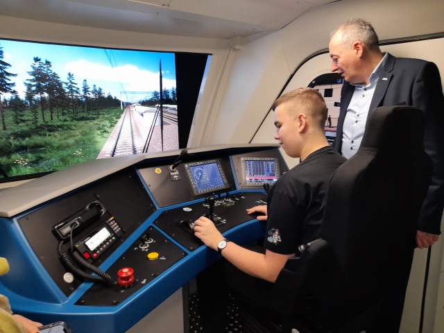 Janek Gawroński ćwiczy na symulatorze pociągu w realistycznie odtworzonej kabinie maszynisty. Przed nim jest pupli i ekran pokazujący renderowaną trasę pojazdu. Nad nim nachyla się doswiadczony maszynista, który udziela mu rad. 