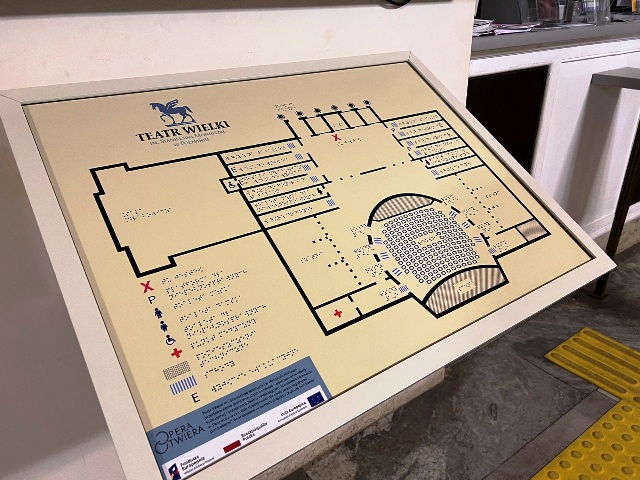 Na zdjęciu widnieje złota tablica w białej ramce (ujęcie tablicy pod kątem około 30 stopni), na której znajduje się plan widowni Teatru Wielkiego w Poznaniu. Opisy wszystkich oznaczonych symbolami miejsc są opisane alfabetem Braille