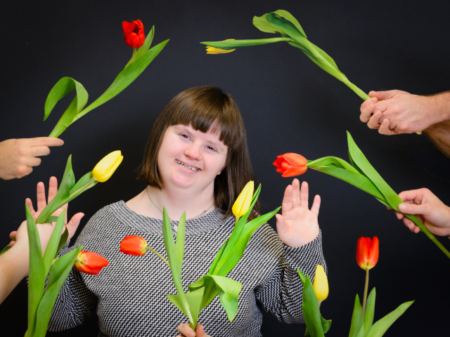 Na środku kobieta z Zespołem Downa, zarówno po lewej i prawej stronie widoczne są dłonie, wręczające tulipany. 
