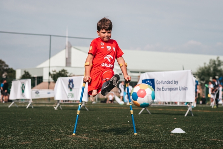 Zawody ampfutbolu. Młody chłopiec opiera się na kulach. Widać, że nie ma jednej nogi. Przygotowuje się do kopnięcia piłki.