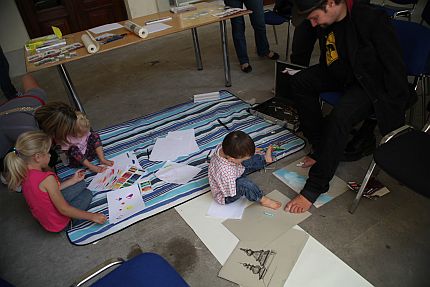 Z prawej siedzi na krześle Stanisław Kmiecik, malując stopami. Na podłodze siedzą dzieci, niektóre z nich także malują stopami