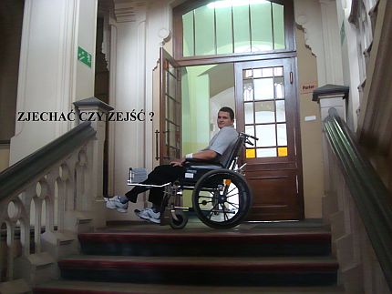 Młody mężczyzna na wózku w zabytkowym wnętrzu, przed nim kamienne schody. Napis: zjechać czy zejść?